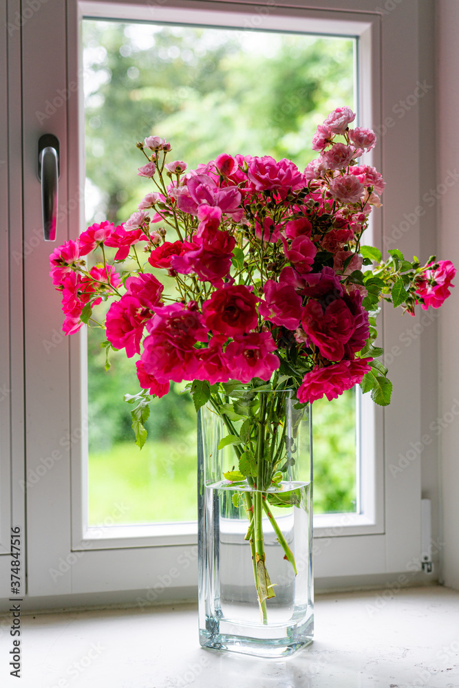 Fototapeta premium Rosenstrauß mit roten Rosen in Glasvase, auf Fensterbrett mit Blick hinaus in einen weiten Garten