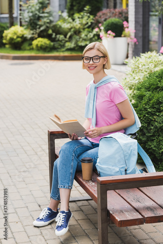 blonde student in glasses holding book while sitting on bench