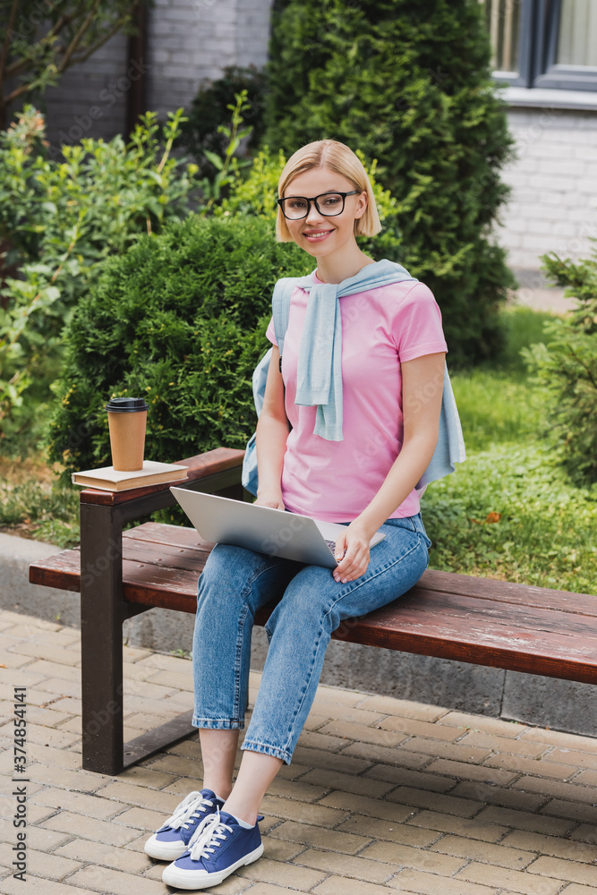 Fototapeta premium blonde student in glasses using laptop near book and paper cup while sitting on bench