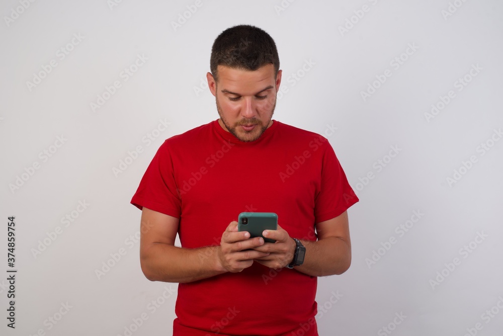 Portrait of focused Young handsome Cucasian man wearing red shirt standing against white background use smartphone reading social media news, or important e-mail