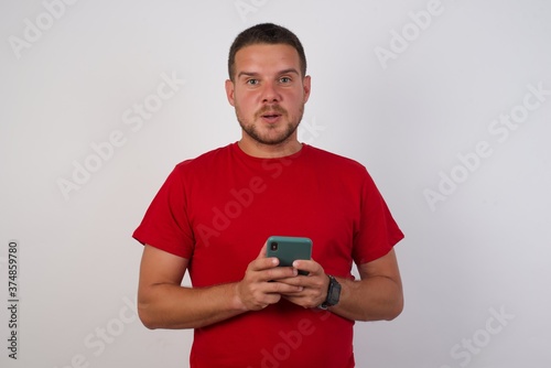 Shocked Young handsome Cucasian man wearing red shirt standing against white background. opens mouth hold phone reading advert unbelievable big shopping prices © Roquillo