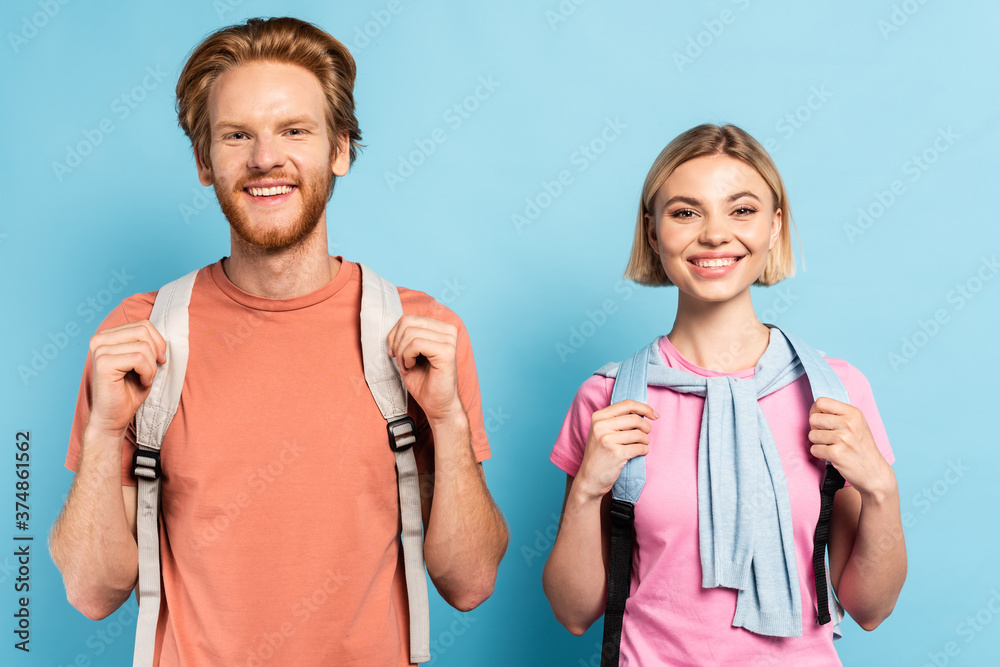 blonde and redhead students with backpacks looking at camera on blue blonde and redhead students with backpacks looking at camera on blue