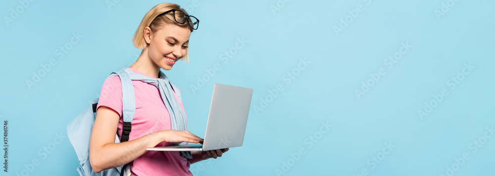 panoramic concept of young blonde student with backpack using laptop isolated on blue - obrazy, fototapety, plakaty 