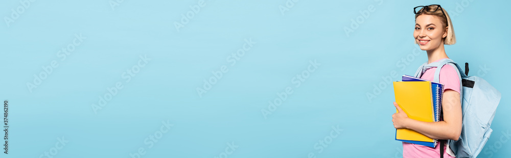 horizontal image of young blonde student with backpack holding notebooks on blue horizontal image of young blonde student with backpack holding notebooks on blue