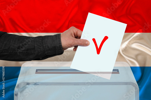 male voter drops a ballot in a transparent ballot box against the background of the Luxembourg national flag, concept of state elections, referendum