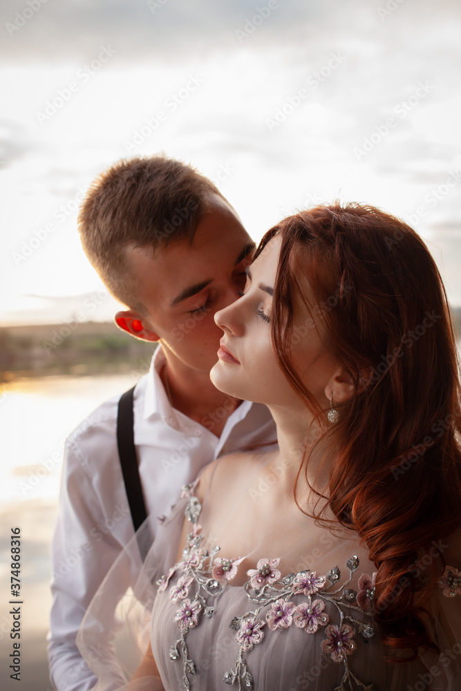 A beautiful wedding couple on the riverbank at sunset. A woman in a gray dress with flowers, a man in a suit and suspenders hug each other, love. Lovers on a walk