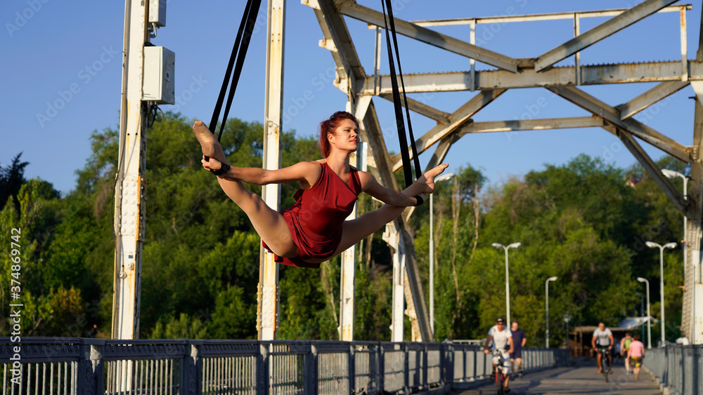 Circus performer on aerial straps performs an aerial act at dawn. flexible, athletic, aerial gymnast in city on metal trusses of bridge engaged in aerial gymnastics. urban acrobatic street show. split Circus performer on aerial straps performs an aerial act at dawn. flexible, athletic, aerial gymnast in city on metal trusses of bridge engaged in aerial gymnastics. urban acrobatic street show. split