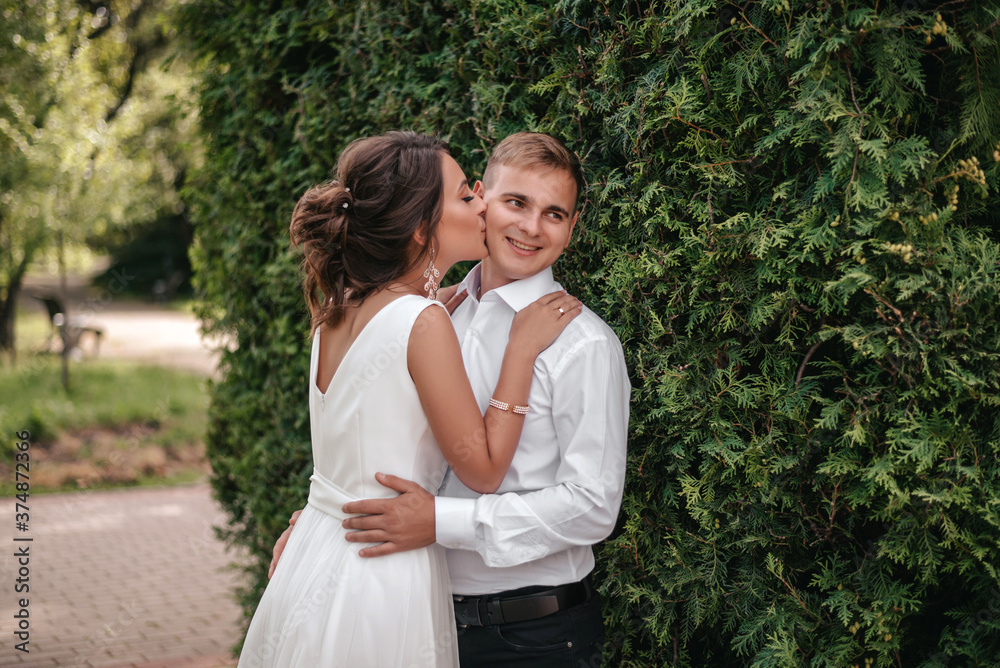Bride kissing on the groom's cheek. Bride wearing white wedding dress The groom wears a suit On their wedding day Evening time, warm sunset light. Bride has hair blond, blue eyes. Beautiful makeup Bride kissing on the groom's cheek. Bride wearing white wedding dress The groom wears a suit On their wedding day Evening time, warm sunset light. Bride has hair blond, blue eyes. Beautiful makeup