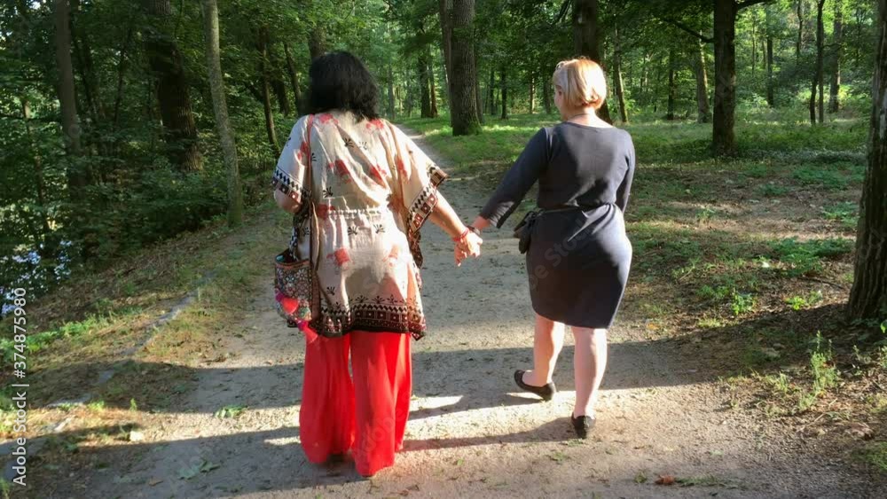Two mature adult women walking in park. On sunny summer day. They kiss each other. From back view. Medium plan. Two mature adult women walking in park. On sunny summer day. They kiss each other. From back view. Medium plan.