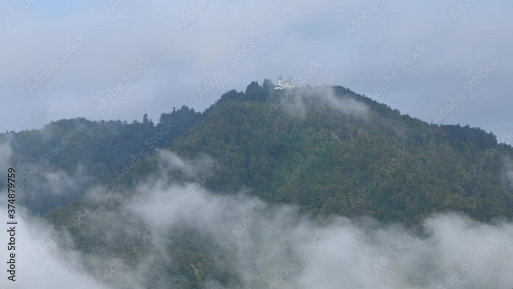 Time lapse low inversion clouds rolling under hill covered with trees. Small catholic church on hilltop Jost in Slovenia. Fog in the valley on a sunny morning. Zoom in Time lapse low inversion clouds rolling under hill covered with trees. Small catholic church on hilltop Jost in Slovenia. Fog in the valley on a sunny morning. Zoom in