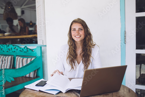 Happy young female freelancer reading book and sitting at table in veranda