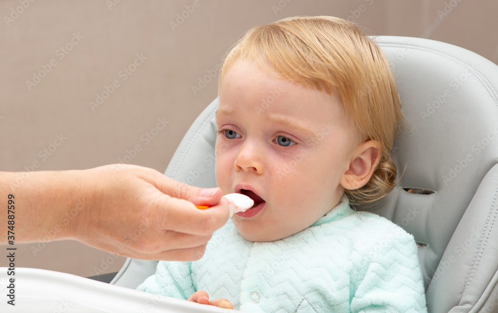 A mother feeds her son in a special baby feeding chair with a small spoon. The mother gives the child food with a child's spoon. Happy baby eats with an appetite. A mother feeds her son in a special baby feeding chair with a small spoon. The mother gives the child food with a child's spoon. Happy baby eats with an appetite.