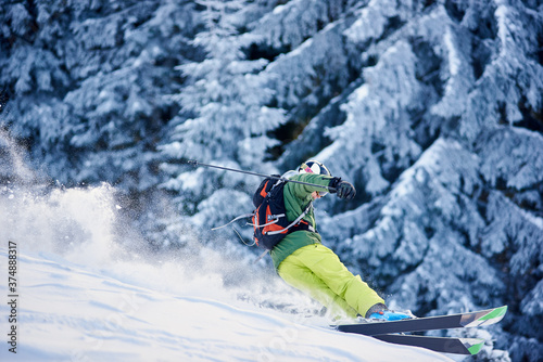 Skier with backpack doing freeride-descent on snow-covered slope in white snow powder blizzard. Dangerous maneuvering on mountain wooded downhill. Picturesque forest scenery on blurred background.
