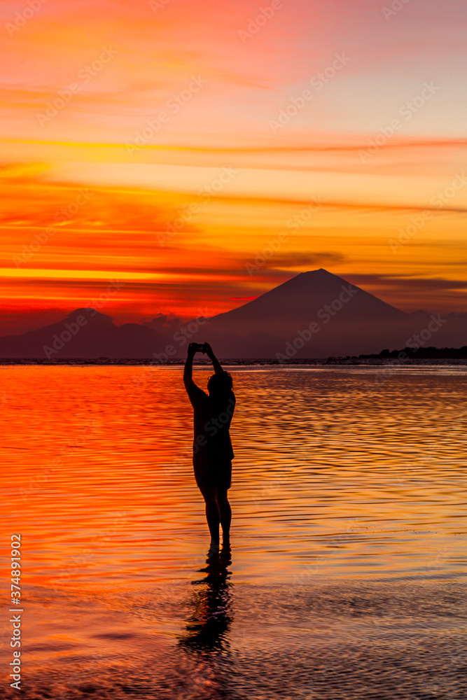 Silhouette of a person taking a photo of the sunset with a volcano in the distance across the ocean