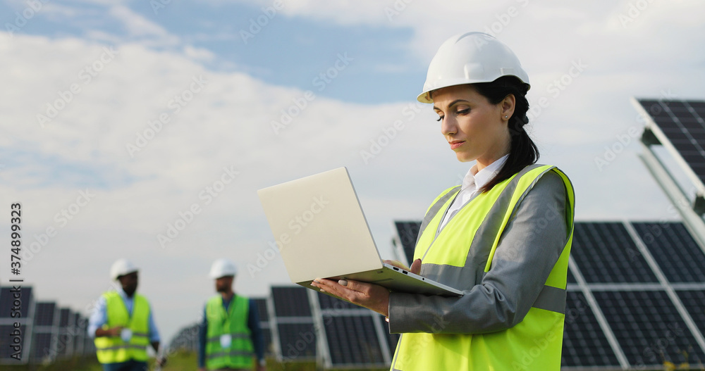 Portrait of electrician engineer in safety helmet and uniform using laptop checking solar panels. Female technician at solar station. Portrait of electrician engineer in safety helmet and uniform using laptop checking solar panels. Female technician at solar station.