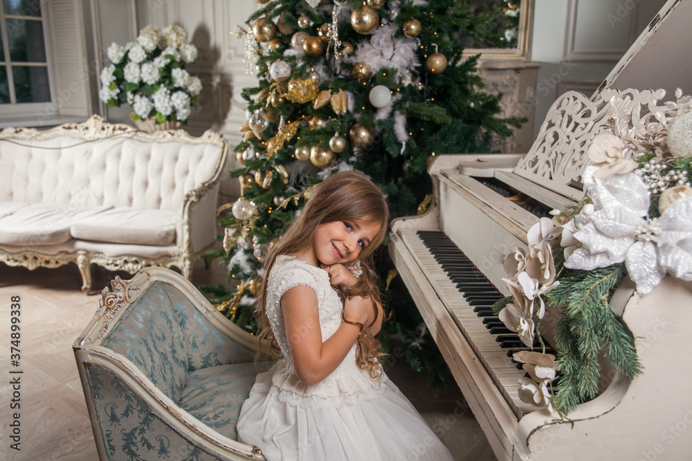 Merry Christmas Happy Holidays concept. Cute little girl in white dress playing on piano against the background of a decorated Christmas tree. Festive New Year home luxury classic interior