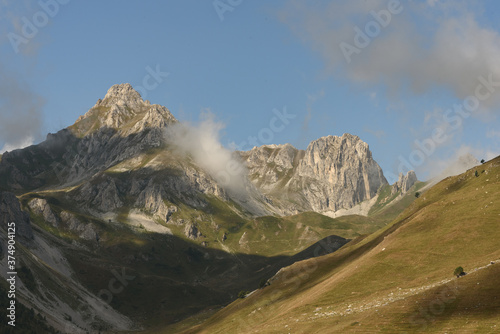 landscape of the mountain peaks of the Alps, rugged and rocky mountains, part of the French Alps mountain range. The sky between clear blue, is covered with white clouds, the valley and grass