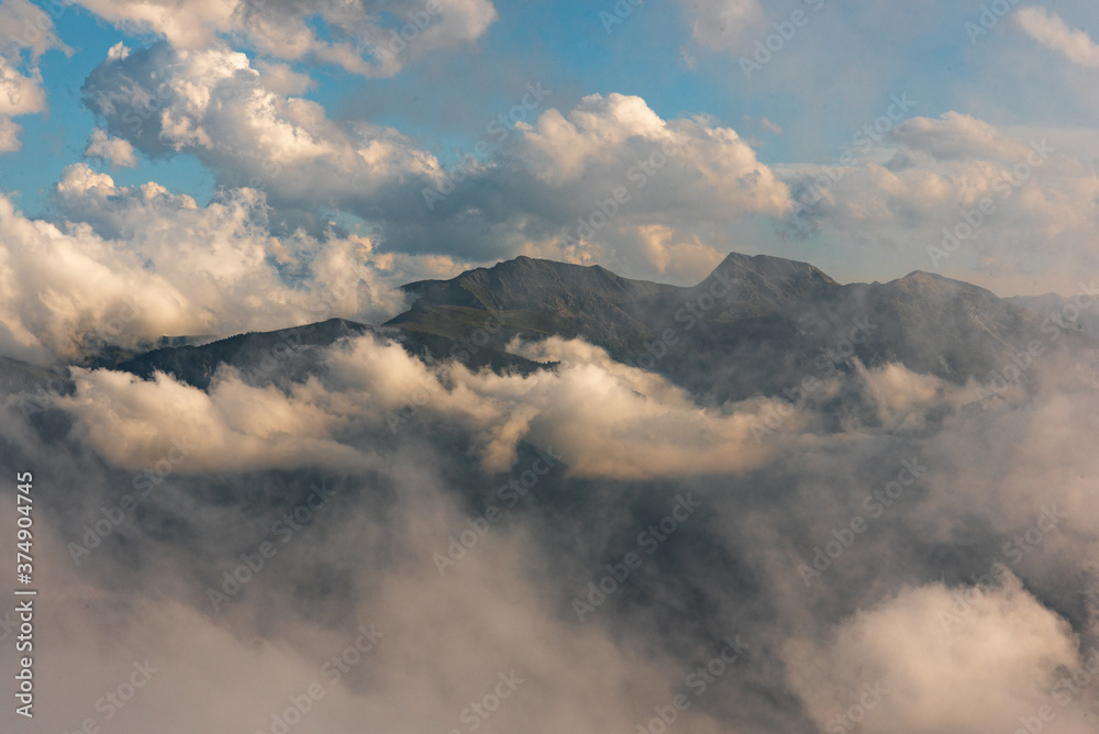 Obraz premium Rocky mountain peak, which rises in the middle of the prairie, in the Alps, between the grass on the slopes of the neighboring mountains and the cloudy sky with small blue glades. Gray rock peak.
