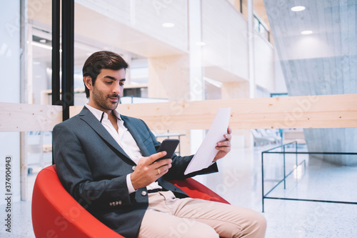 Calm stylish businessman with papers interacting with smartphone in lobby of modern office building