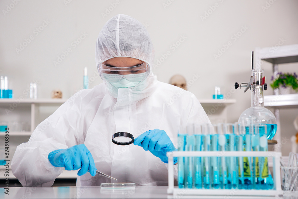 Male Scientist in PPE Suit working in Lab while Using Magnifying glass Checking Sample in Sampling Plate. Blue Tone. SARS-CoV-2 , Covid-19 THEME. Male Scientist in PPE Suit working in Lab while Using Magnifying glass Checking Sample in Sampling Plate. Blue Tone. SARS-CoV-2 , Covid-19 THEME.