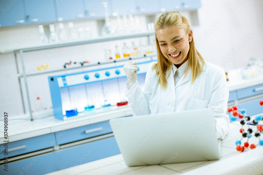 Happy female researcher in white lab coat using laptop while working in the laboratory - obrazy, fototapety, plakaty 