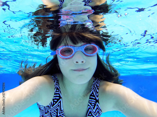 Underwater photo of girl swimming in a pool. Child swims underwater and looking at camera close up while wearing goggles. Clear blue water, reflections, light and water surface visible.
