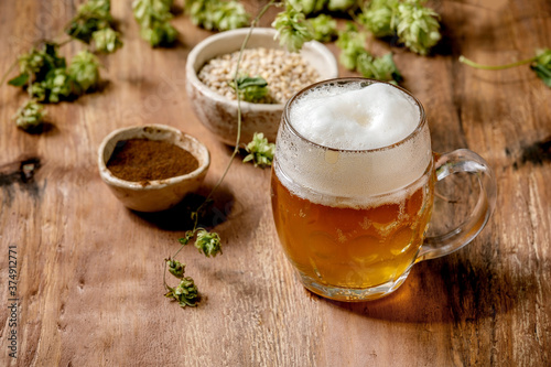 Classic glass mug of fresh cold foamy lager beer with green hop cones, wheat grain and red fermented malt in ceramic bowls behind over wooden texture background. Copy space