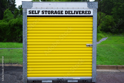Portable self-storage unit on a street or parking lot.  Yellow door and locking mechanism visible. Metal storage shed delivered to a location.