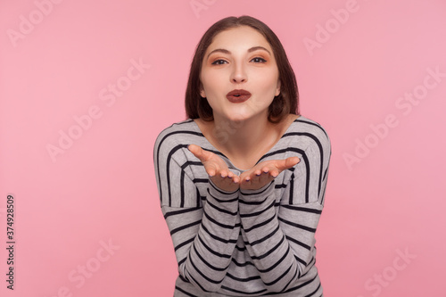 Love you  Portrait of sensual attractive woman in striped sweatshirt sending air kiss over palms  expressing fondness  romantic feelings and flirt. indoor studio shot isolated on pink background