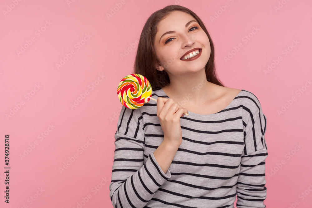 Portrait of cheerful woman in striped sweatshirt holding sweet round rainbow candy lollipop and looking at camera with happy childish expression. indoor studio shot isolated on pink background Portrait of cheerful woman in striped sweatshirt holding sweet round rainbow candy lollipop and looking at camera with happy childish expression. indoor studio shot isolated on pink background