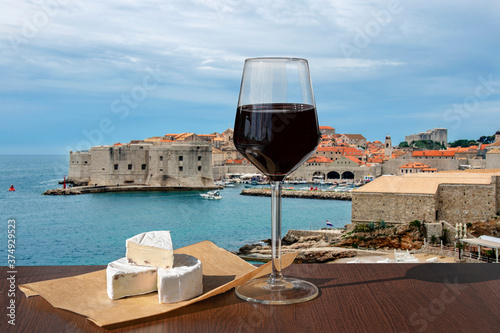 Fototapeta Naklejka Na Ścianę i Meble -  Glass of red wine with brie cheese against Dubrovnik city center background. Vacation concept. Beach and city wall in historic center of Dubrovnik, Croatia