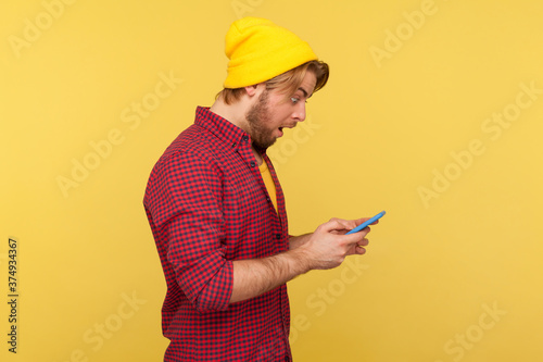 Side view amazed guy in hipster beanie hat and shirt looking surprised at cell phone, reading shocking message, chatting in social network, astonished by mobile app. indoor studio shot isolated
