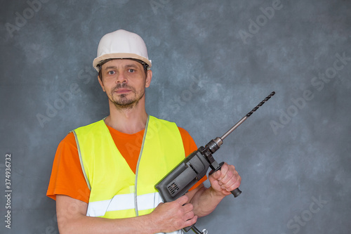 A 40-year-old European man holds an electric drill in his hands. A worker in a helmet and a vest with a tool in his hands. © Sergei