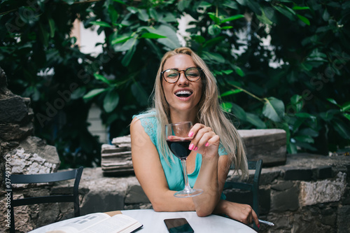 Portrait of joyful female holding glass with red wine enjoying weekend for drinking alcohol and rest in street cafeteria, happy woman in eyeglasses for vision correction laughing photo