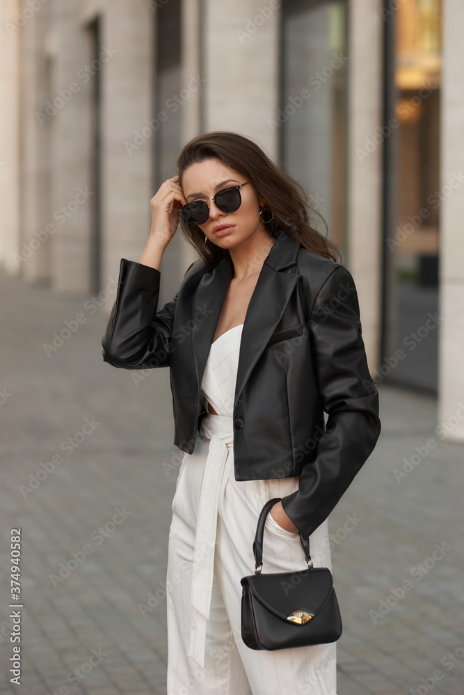 Young stylish woman in white overall, sneakers, black leather jacket and sunglasses holding small handbag and standing at city street. Fashion style outdoor portrait