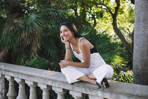 Smiling brunette hipster girl in trendy wear laughing outdoors satisfied with free time vacation in park, happy pretty 20s woman in stylish outfit sitting outdoors enjoying trip weekends in summer #374940904