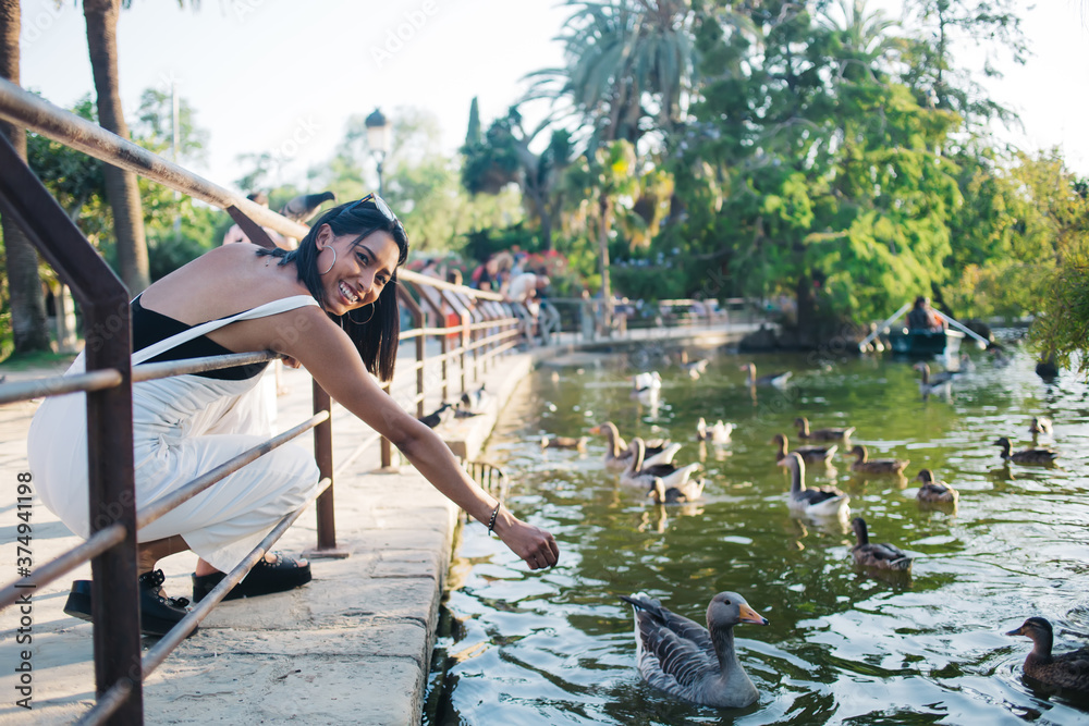 Happy caucasian 20s woman looking at camera giving food to domestic birds on lake in city park recreating on vacations, smiling female traveler feeding ducks and swans having fun on weekends in town