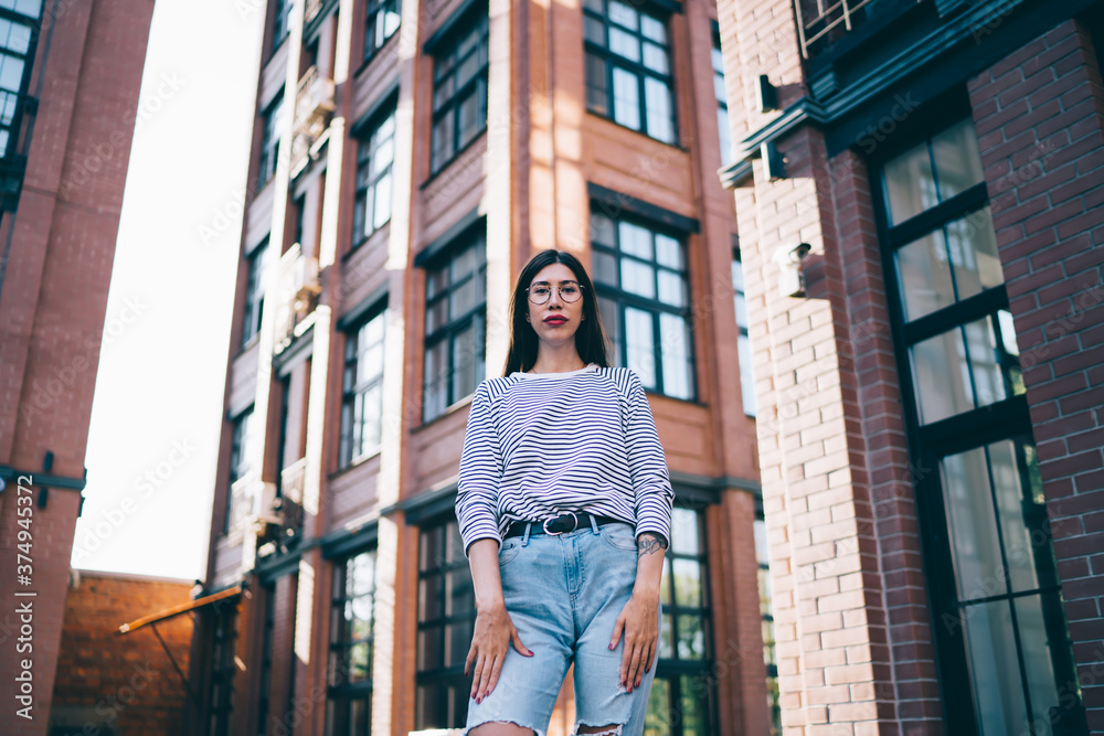 Half length portrait of trendy dressed female teenager in stylish optical spectacles for provide eyes protection standing at city street with facade buildings on background and looking at camera
