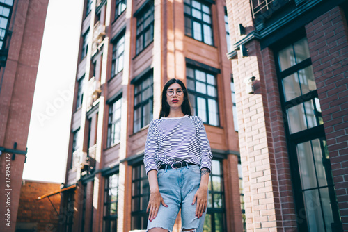 Half length portrait of trendy dressed female teenager in stylish optical spectacles for provide eyes protection standing at city street with facade buildings on background and looking at camera