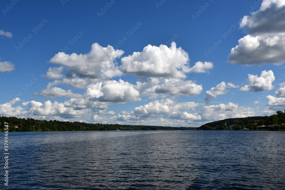 Blue sky with beautiful white clouds over a wide river. Ripples on the surface of the water. Low cloud. Green forest on the banks.