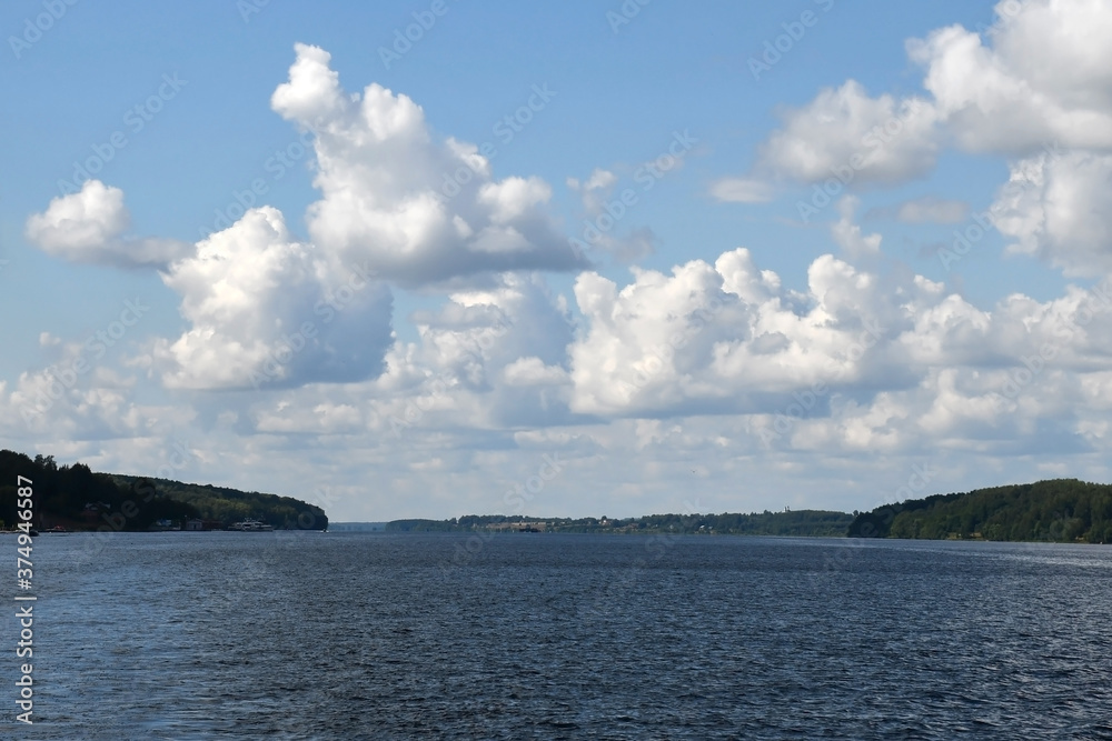 Blue sky with beautiful white clouds over a wide river. Ripples on the surface of the water. Low cloud. Green forest on the banks. Blue sky with beautiful white clouds over a wide river. Ripples on the surface of the water. Low cloud. Green forest on the banks.
