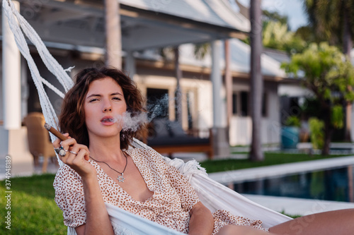 Fotografie Young european woman smoking cigar lying on hammock outside tropical luxury villa hotel, sunset natural light