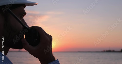 A seaport worker puts on a white helmet and talks on the radio. A seaport worker manages logistics by radio communication  on the background of the sea early in the morning.