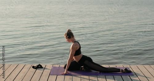 Young woman is training outdoors. Sports, yoga, stretching, wellness concept. Female is practicing yoga doing pigeon pose on sport mat on river pier listening music in headphones, side view.