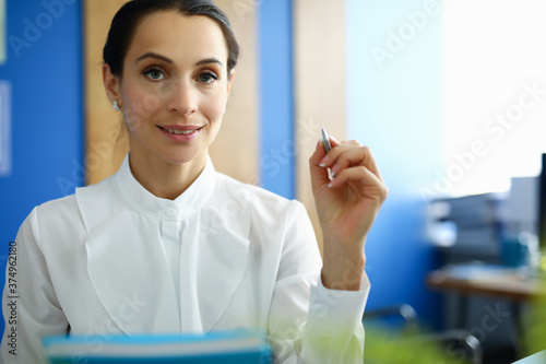 Beautiful businesswoman in white shirt hold pen with document in her hand and smile. In background is workplace in office.
