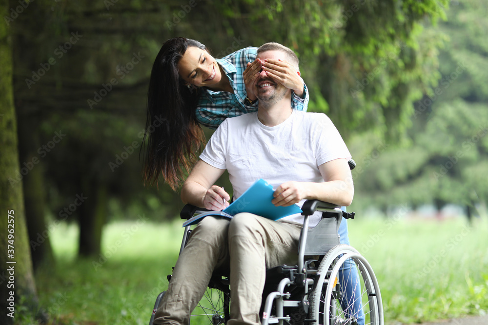 Happy woman stand behind and cover man eye with her palm. Cheerful man sit in wheelchair and hold notebook with pen in his hand.