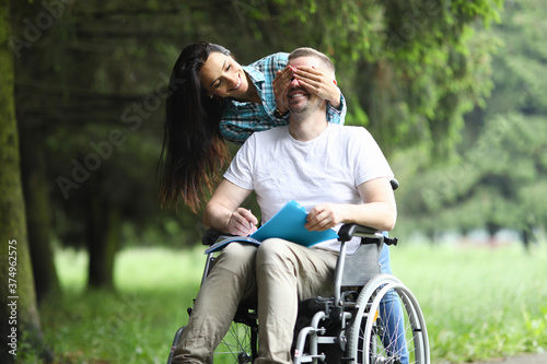 Happy woman stand behind and cover man eye with her palm. Cheerful man sit in wheelchair and hold notebook with pen in his hand.