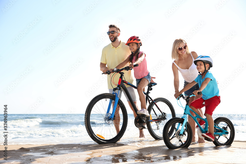 Fototapeta premium Happy parents teaching children to ride bicycles on sandy beach near sea