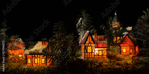 Panoramic scene of miniature Christmas countryside with lit tiny ceramic houses. Realistic homemade Xmas model of night village with trees, fake snow on roofs and Christian church on black background.