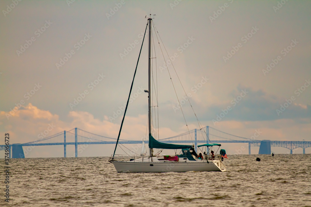 Image of a sailboat moving across Chesapeake Bay with the silhouette of the famous Bay Bridge in the background. There are people on the boat who are enjoying the sunset over the bay,.